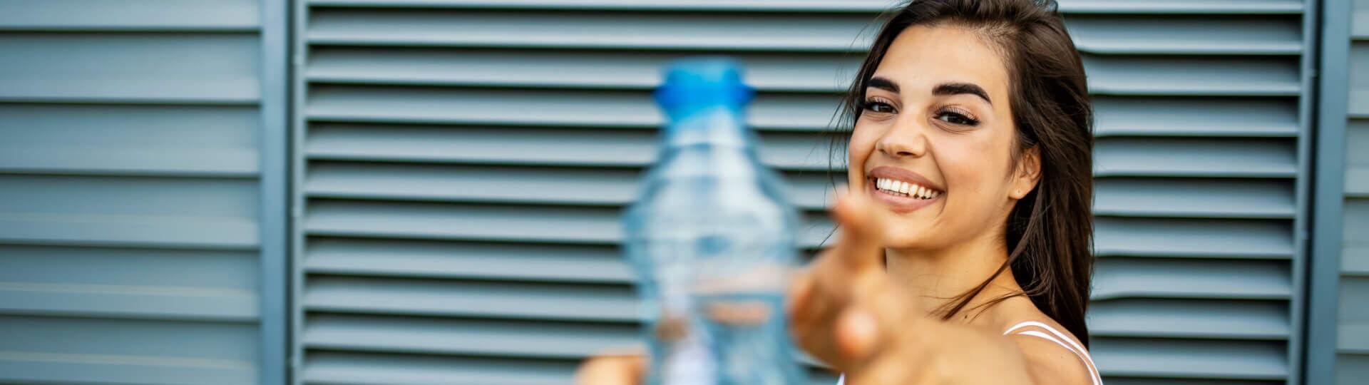 Eine Frau im Sportoutfit und Yogamatte in der Hand wird eine Wasserflasche gereicht