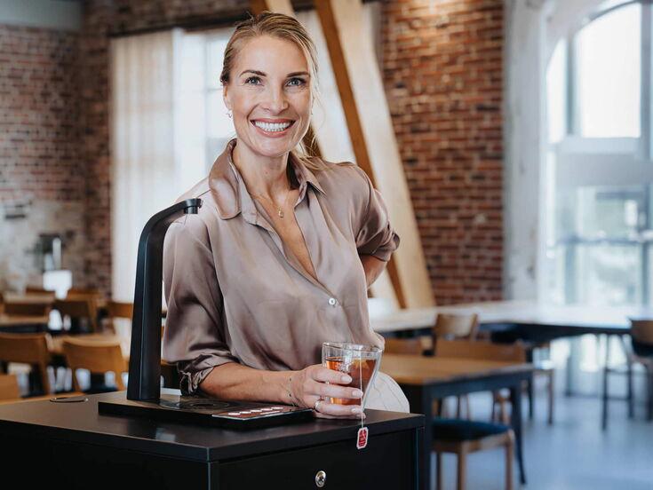 Eine Frau lächelt, während sie mit einem Teeglas in der Hand neben einem eleganten Wasserzapfhahn steht. Im Hintergrund sind Holzmöbel sichtbar.